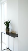 Minimalist entryway with black console table, snake plant in dark pot, and gold welcome sign against bright white walls near window.