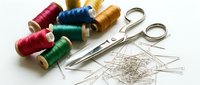 Colorful thread spools in red, pink, blue, gold and green arranged with metal scissors and scattered sewing needles on white background for crafting and tailoring projects.