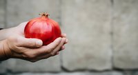 Hand holding fresh red pomegranate fruit against blurred concrete wall background, suitable for healthy eating, nutrition, and food photography projects.