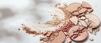 Broken makeup powder compacts with scattered cosmetic dust creating artistic beauty product composition on white background for skincare advertising.