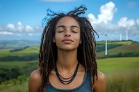 Young African American woman with dreadlocks and closed eyes enjoying peaceful moment outdoors against green hills and wind turbines background on sunny day.