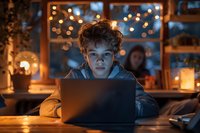 Young boy illuminated by laptop screen in dark room with bokeh lights and candles in background, creating moody atmospheric evening scene at home.