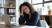 Stressed businesswoman with curly hair holding head in hands at office desk with laptop experiencing work burnout and mental fatigue in modern workplace.