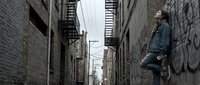 Young man standing in urban alley with brick walls and fire escapes, contemplating city life in moody atmospheric street scene.