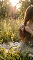 Young woman braiding long hair in golden sunset meadow with wildflowers and warm sunlight filtering through trees creating peaceful natural atmosphere.