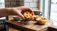 Hand holding freshly baked Portuguese custard tart with caramelized top on wooden cutting board in modern kitchen setting for bakery and cooking content.