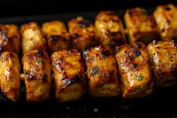 Grilled chicken skewers with golden-brown caramelized glaze and char marks on dark background, close-up macro shot showing juicy meat texture and marinade details.