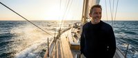 Man in black sweater standing on sailing yacht deck during golden hour with ocean waves and rigging visible, suitable for travel, adventure, and lifestyle content.
