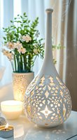 Elegant white ceramic vase with intricate cutout pattern glowing from interior light, alongside flowers and candles creating cozy atmospheric home decor setting.