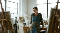 Female artist of mixed ethnicity standing in bright studio surrounded by easels, canvases and art supplies, wearing apron and plaid shirt, contemplating creative work.