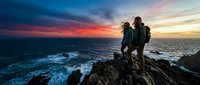 Couple standing on rocky cliff overlooking ocean at sunset, dramatic sky with orange and blue tones, waves crashing below, travel and adventure concept.