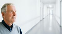 Senior man with gray hair and Caucasian ethnicity wearing casual blue shirt, standing in bright hospital or medical facility corridor, looking thoughtfully to the side in healthcare setting.