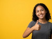 Young woman with long curly dark hair and mixed ethnicity, wearing gray t-shirt, giving thumbs up gesture with bright smile against yellow background for approval and success concepts.