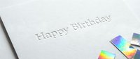 Happy Birthday embossed text on white textured card with holographic confetti, elegant minimalist greeting card design for celebration.