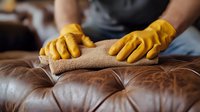Person wearing yellow rubber gloves cleaning and conditioning brown leather sofa with microfiber cloth, close up view of furniture maintenance and care at home.