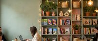 Modern cozy cafe interior with wooden bookshelves, plants and warm lighting. Young woman working on laptop in comfortable reading space for remote work.