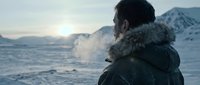 Young man with dark hair and beard wearing heavy winter clothing with fur-lined hood, standing in snowy arctic landscape at sunset, gazing toward frozen mountains and icy terrain.