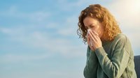 Woman with curly reddish-brown hair, appearing to be of Caucasian ethnicity, wearing a sage green cardigan, sneezing or coughing into her tissue outdoors against a blue sky. Suitable for health, wellness, and seasonal illness content.