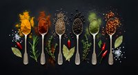 Colorful spices and herbs arranged on wooden spoons against dark background. Culinary ingredients including turmeric, paprika, seeds for cooking and seasoning.