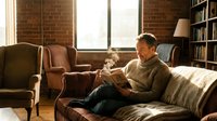 Middle-aged Caucasian man in cozy sweater reading book on sofa in warm living room with brick walls, bookshelves and natural light streaming through windows.
