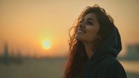 Young woman with curly hair in hoodie enjoying peaceful sunset moment by water with warm golden light and serene expression for wellness lifestyle content.