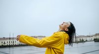 Woman in yellow raincoat with arms outstretched facing water, expressing freedom and joy during rainy weather for lifestyle and wellness content.