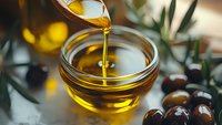 Golden olive oil pouring from wooden spoon into glass bowl with fresh olives and green leaves on rustic kitchen table for cooking and culinary use.