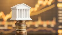 White miniature bank building model on stack of golden coins with blurred financial charts background representing banking investment growth concept.