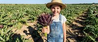 Young female farmer in overalls and hat holding fresh pineapple in tropical plantation field during harvest season under sunny sky.