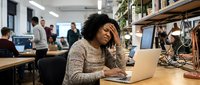 Stressed African American businesswoman working on laptop in busy office environment, experiencing work pressure and fatigue during demanding workday.