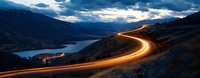 Dramatic light trails from vehicle headlights curve along mountain road at dusk, overlooking serene lake valley with scattered city lights below and moody clouded sky.