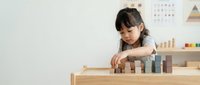 Young Asian girl playing with wooden building blocks at table in bright educational playroom developing fine motor skills and creativity.