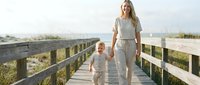 Young blonde mother and toddler daughter walking hand in hand on wooden boardwalk during golden hour, wearing matching casual outfits for family lifestyle content.