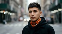 Young Hispanic man in black hoodie and orange bandana standing on urban city street with blurred traffic lights and buildings in background.
