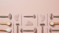 Rose quartz facial massage tools and metal beauty instruments arranged in organized pattern on pink background for skincare routine.