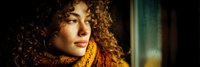 Young woman with curly hair looking thoughtfully out window with warm golden light creating dramatic shadows and highlights on her face.