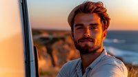 Young Caucasian man with beard and styled hair smiling at sunset near coastline, wearing white shirt, warm golden hour lighting creates atmospheric outdoor portrait.
