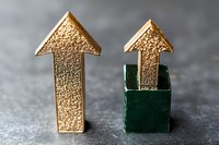 Golden upward arrows with textured surface, one emerging from green cube box on gray concrete background, symbolizing growth and breakthrough in business.