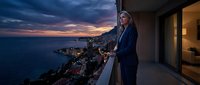 Professional businesswoman in elegant suit standing on luxury balcony overlooking coastal city at sunset, contemplating success and future opportunities.