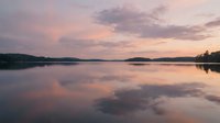 Serene lake at sunset with pink and purple sky reflecting on calm water, surrounded by forested shoreline and distant islands during golden hour.