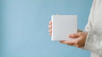 Person with medium skin tone wearing light gray sweater holding blank white box against blue background for product display or packaging mockup.