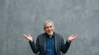 Senior man with white hair and glasses, wearing dark blazer over blue shirt, gesturing with open palms in welcoming pose against gray textured background. Suitable for business, leadership, and corporate communications.