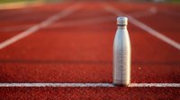 Stainless steel water bottle on red athletic track with white lane markings, suitable for sports, fitness, hydration and wellness marketing.