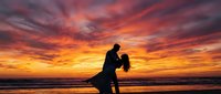 Romantic couple silhouette dancing on beach at sunset with dramatic orange and pink sky clouds reflecting on ocean waves creating intimate moment.