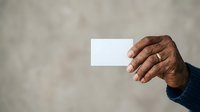 Close-up of mature male hand with dark skin tone holding blank white business card against neutral gray background for corporate branding.