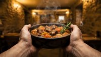 Male hands holding steaming bowl of hearty stew with vegetables and meat in rustic kitchen setting, comfort food preparation and serving concept.