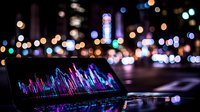 Laptop displaying colorful financial charts and graphs at night with blurred city lights bokeh background, modern business analytics and trading concept.