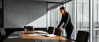 Professional businesswoman in dark suit standing at modern conference table in corporate office with large windows and natural lighting for business presentations.