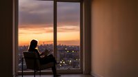 Woman silhouette reading book by large window overlooking city skyline at sunset with warm golden light creating peaceful indoor atmosphere for relaxation.
