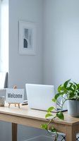 Minimalist home office desk with laptop, welcome sign on easel, and green potted plant against a light gray wall with framed art.